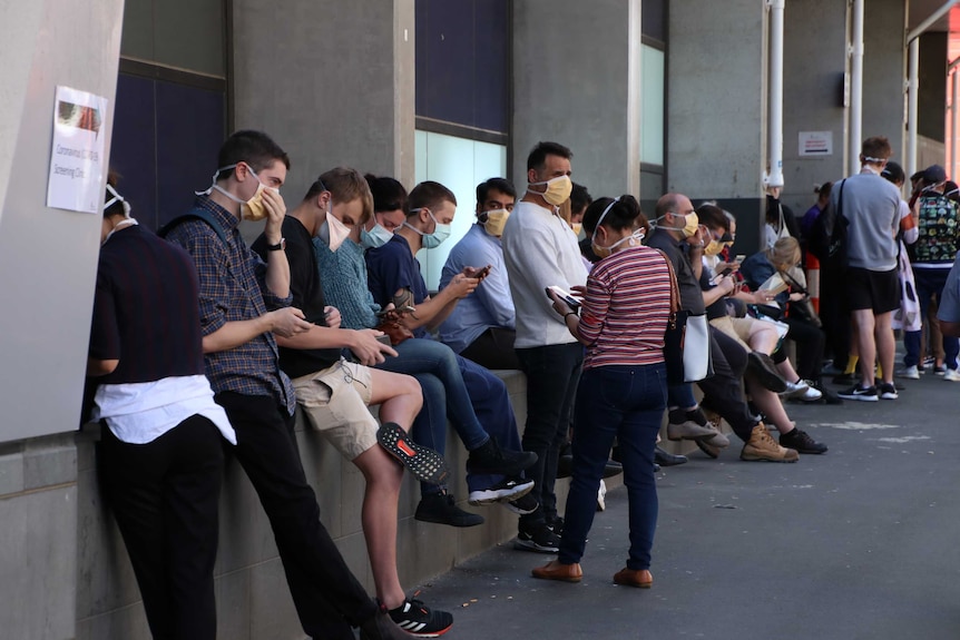 About a dozen people wearing masks sitting outside looking at their phones in a queue.