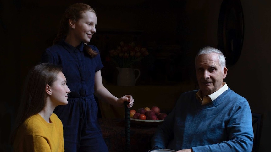 A silver-haired man is seated with two granddaughters near a table with fruits.