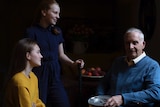 A silver-haired man is seated with two granddaughters near a table with fruits.