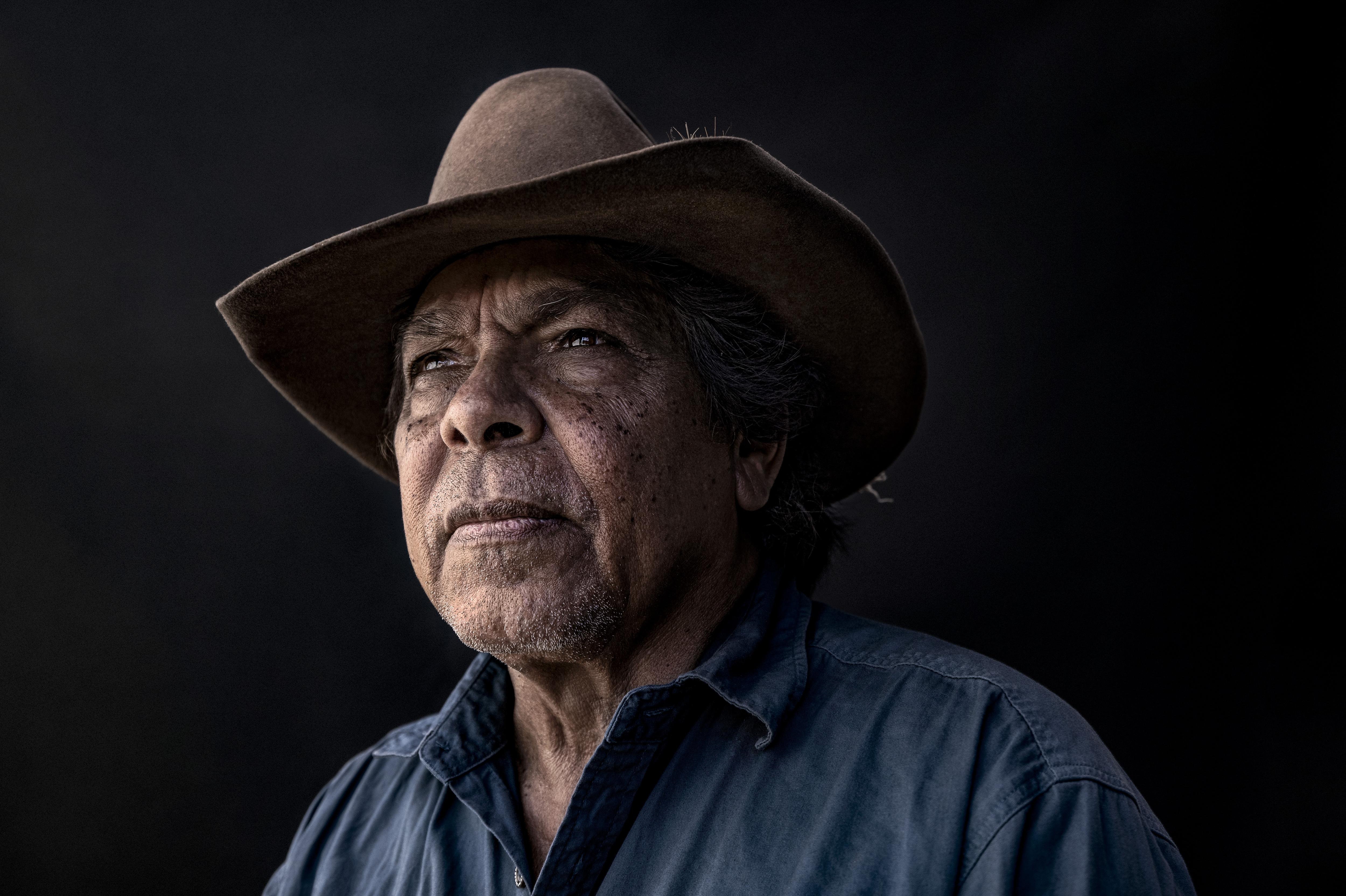 an indigenous man in a brown cowboy hat and dark blue button up shirt looks to the left of the camera