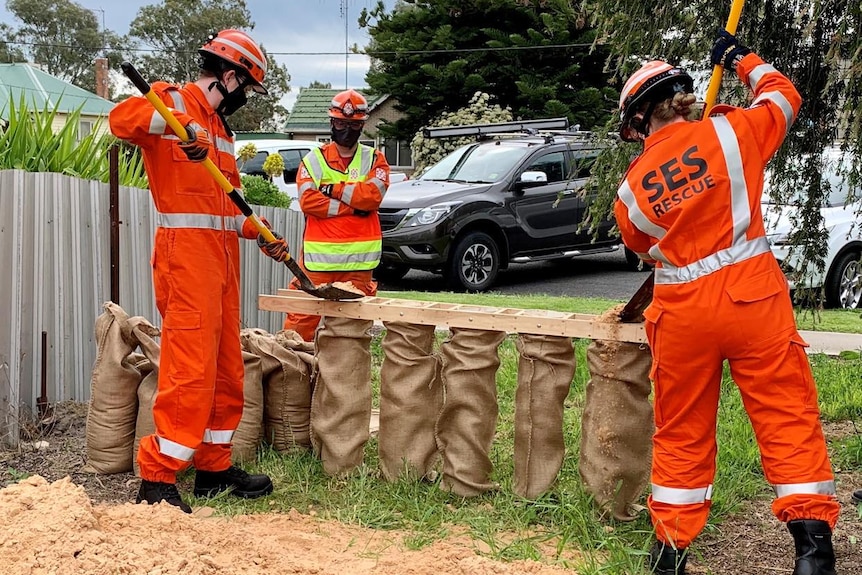 three people in SES uniforms use a shovel to fill bags with sand