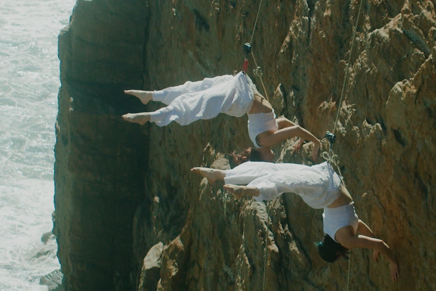 Two women suspended upside down on a cliff face wearing white costumes. 