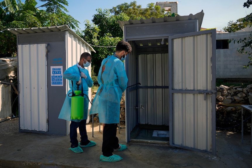 Two workers in protective gear spray a metal outhouse toilet. 