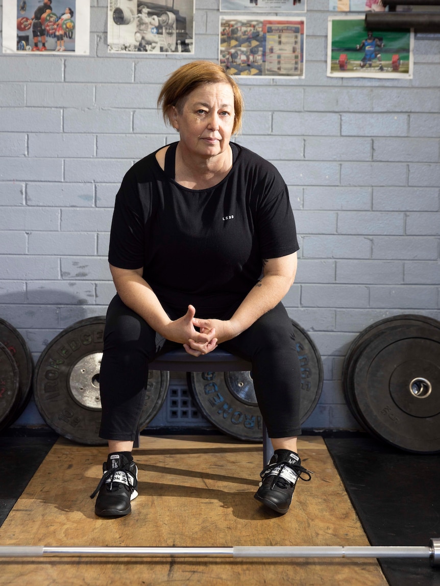 A middle-aged woman sits on a bench in a gym and looks at the camera with a serious expression
