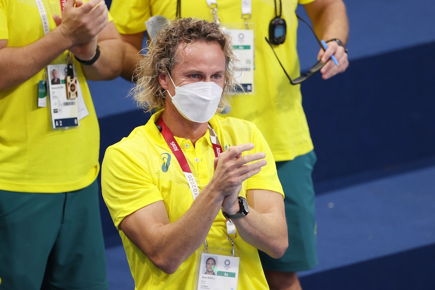 A blonde man wearing a yellow shirt claps his hands