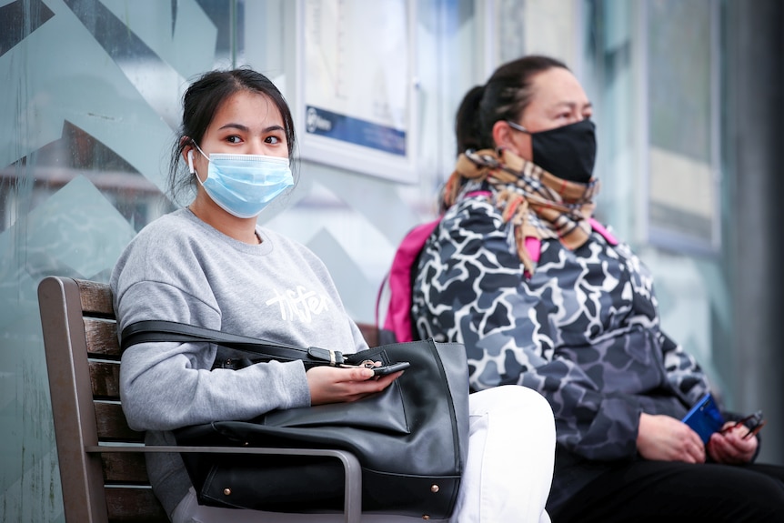 Two women in surgical masks sitting on a bench at a bus stop