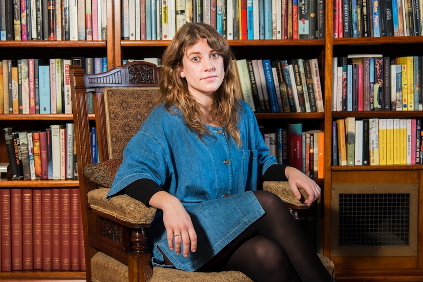 A brunette woman sits in an armchair, in front of stacked bookshelves, looking into the camera with a serious expression