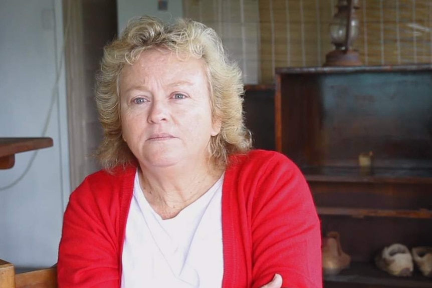 Portrait of an older woman with blue eyes and blonde hair sitting in her kitchen.