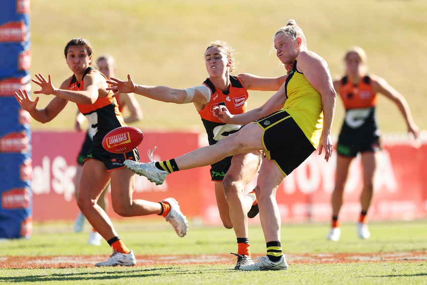 A Richmond AFLW player swings her leg to kick it toward goal as GWS defenders reach out to block.