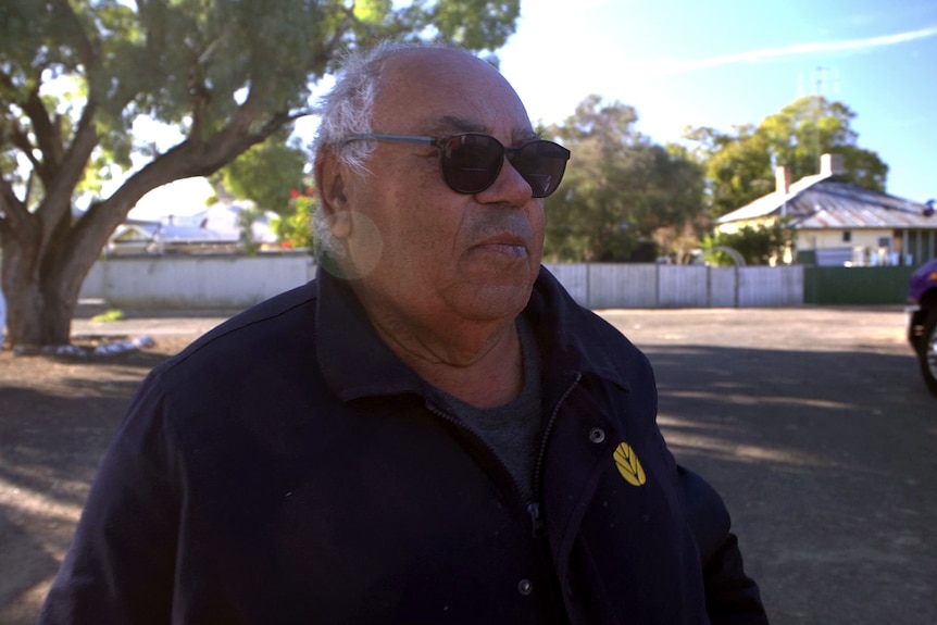 A man with white hair and wearing sunglasses looks out with a car park behind him.