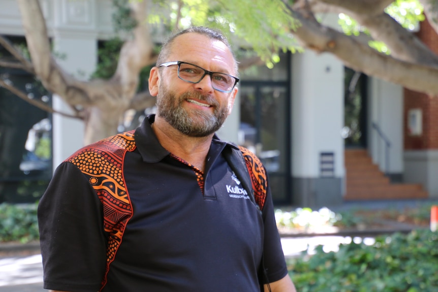 A man with a beard and glasses, wearing a black polo with Aboriginal patters on the shoulders, smiles at the camera in a park.