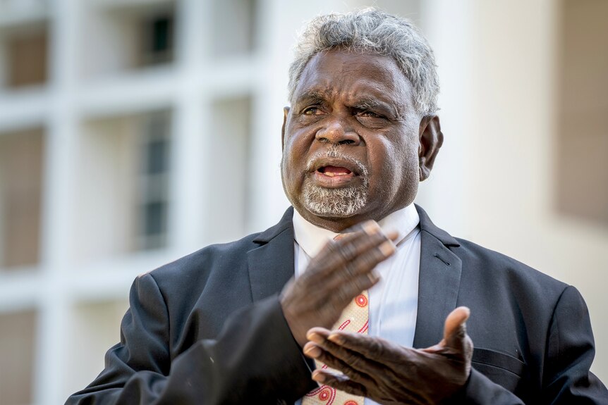 a photo of a man in a suit speaking with a building in the background