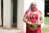 A woman with long white hair wearing a dress holds some leaves in her hands as she looks at the camera.