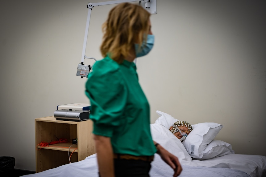 Angela D’Rozario wearing a surgical mask and walking past a clinical-looking bed, where a per sleeps in an electrode cap.