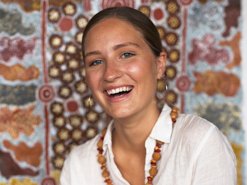 Rona smiles at the camera, wearing a white shirt and a beaded necklace.