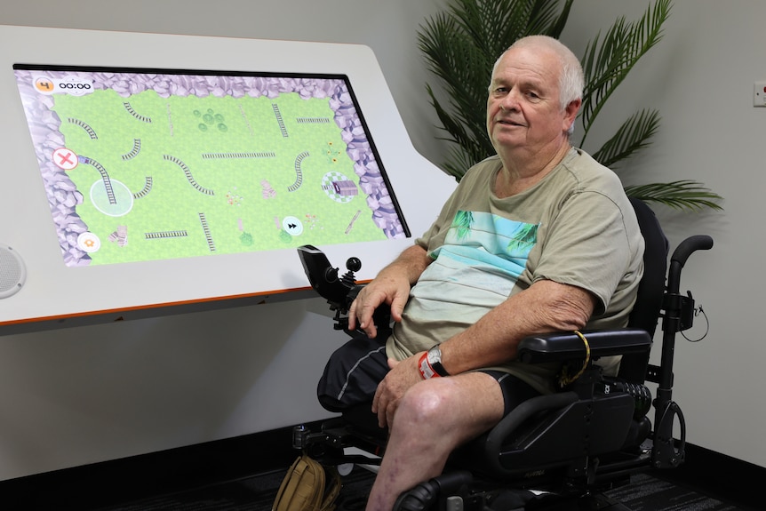 A man in a wheelchair sitting in front of a large computer screen smiling at the camera
