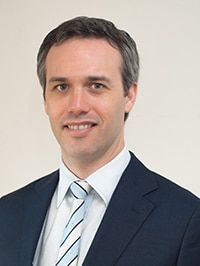 Edward Santow smiles while wearing a suit with a striped tie.