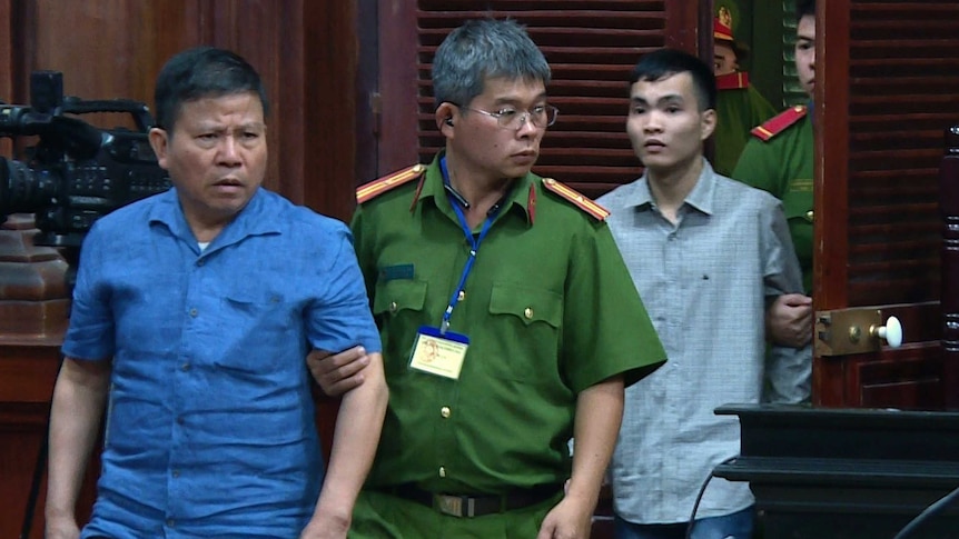 A man in blue with a furrowed brow is escorted by a guard in green into a court room.