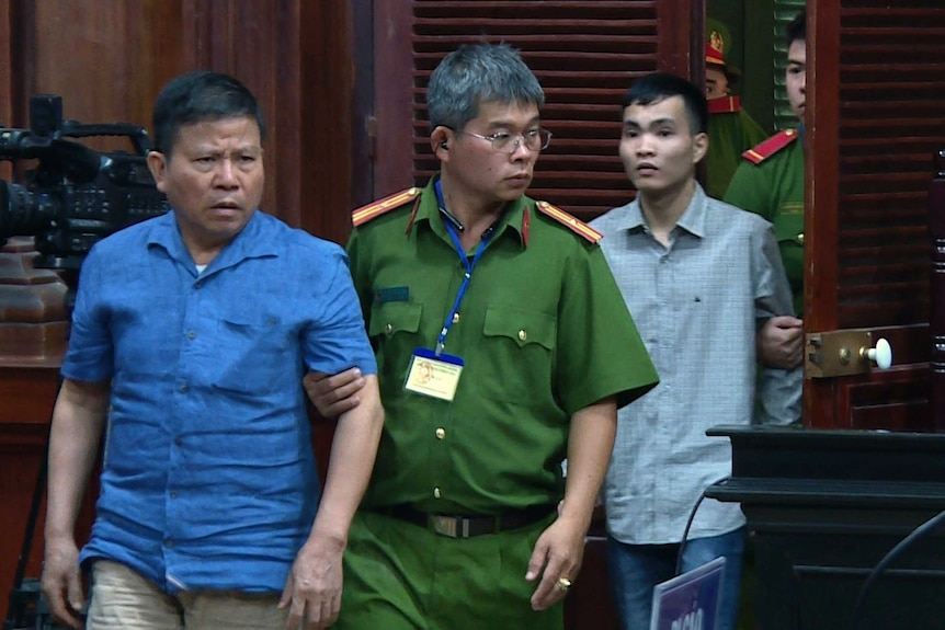 A man in blue with a furrowed brow is escorted by a guard in green into a court room.