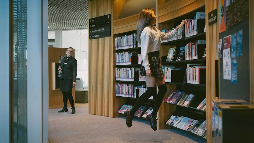 A woman apparently levitates while reaching for a book in a library; another woman looks on