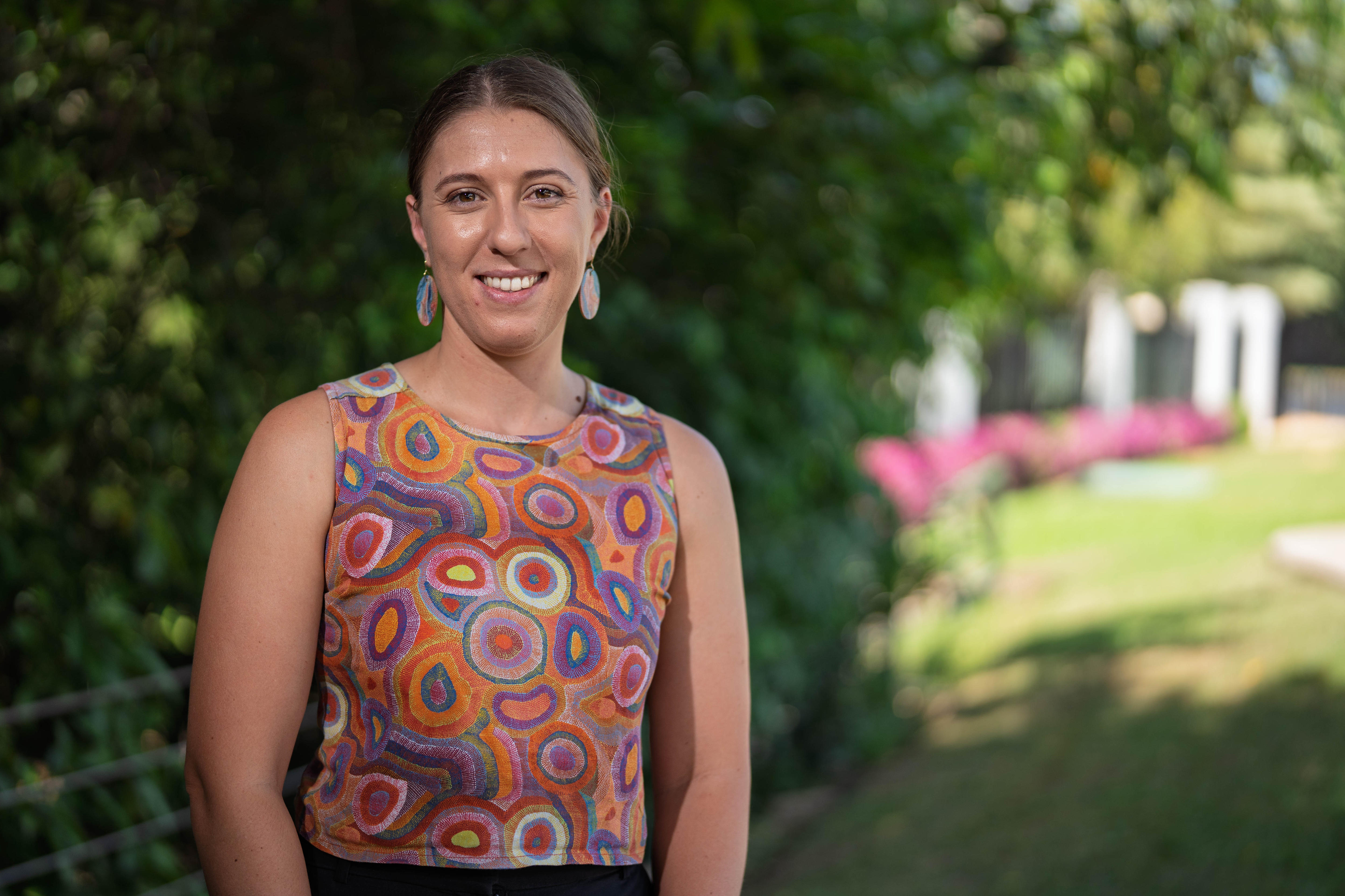 A young woman, Dr Dayna Duncan, smiles at the camera. She is wearing a colourful shirt and standing in a park.