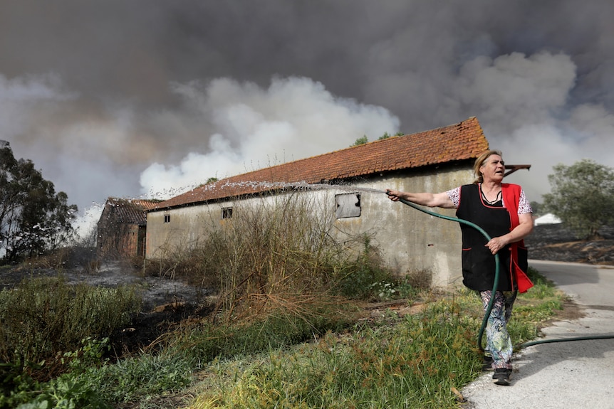 A woman stands with a hose shooting water at a fire. Smoke billows from a house in the background. 