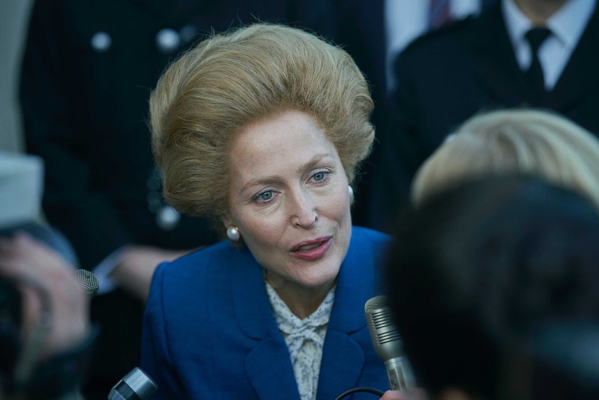 A middle aged woman in a blue suit fronts the press in a scene from The Crown.