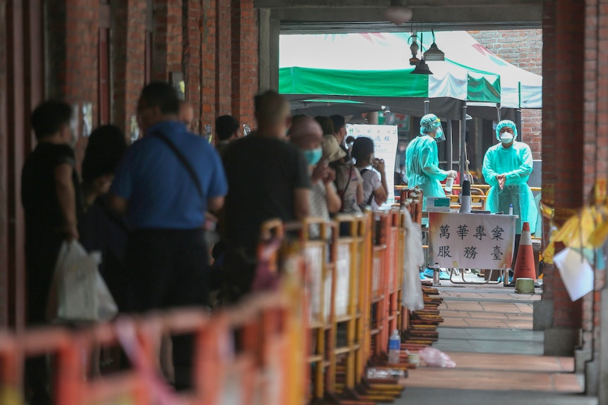 Two health workers in green scrubs and masks stand at the head of a queue of people 