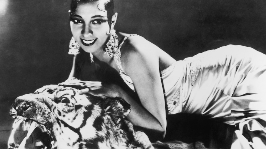 Black and white portrait of a young Josephine Baker with lavish diamond earrings in a sleek gown lying on a rug