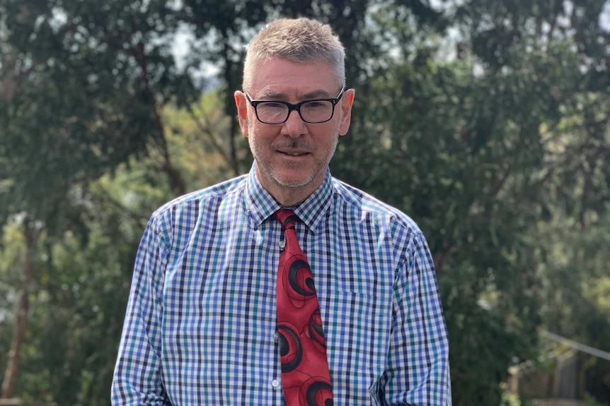 A man with grey hair wearing a shirt and red tie stands outside holding a mask.