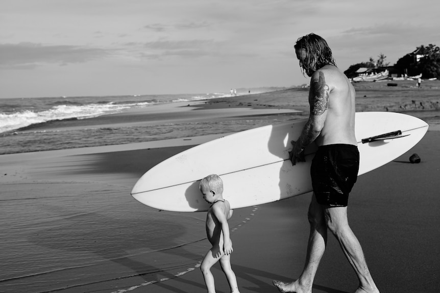 A man walks on the beach holding a surf board, with a young boy walking next to him.