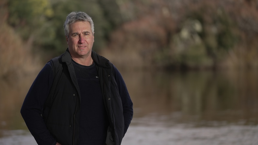 A man standing near a river wearing a navy shirt and vest. 