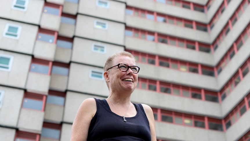 Stef with blonde shirt hair and a blue singlet smiling in front of a high-rise housing commission building.