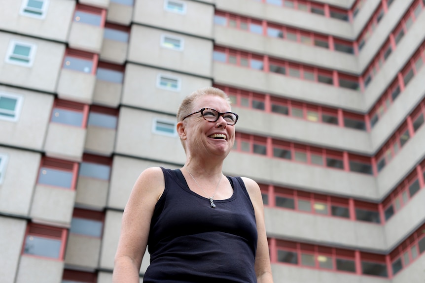 Steph with blonde shirt hair and a blue singlet smiling in front of a high-rise housing commission building.
