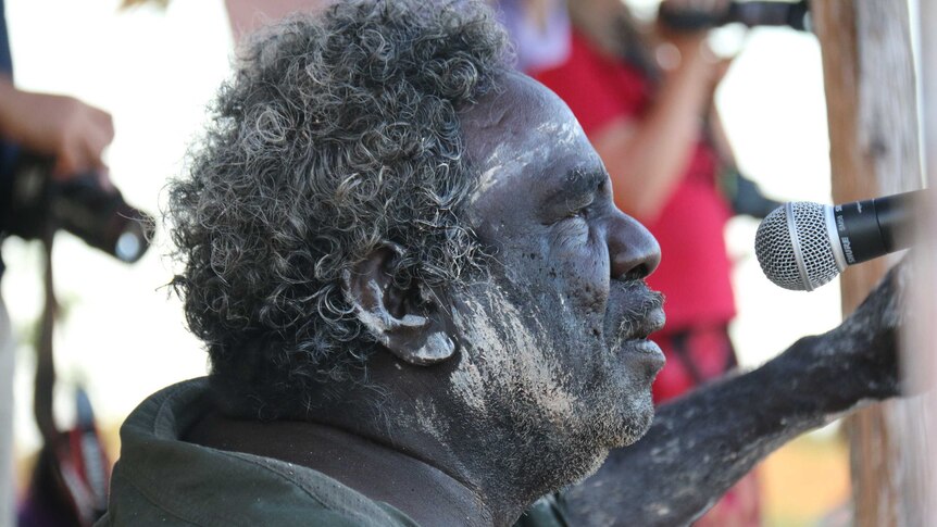 A close-up of Aboriginal man Michael Mungala painted with white mud and singing into a microphone.