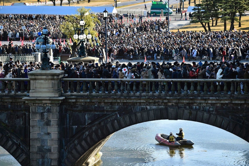 Thousands of people stand in a line on a bridge as a boat paddles underneath
