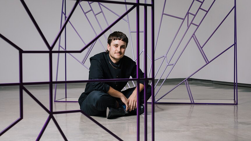 Colour photo of artist Spence Messih sitting on museum floor in front of his sculptures at the Museum of Contemporary Art.