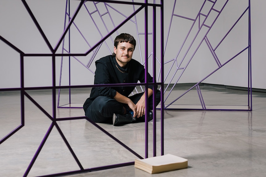 Colour photo of artist Spence Messih sitting on museum floor in front of his sculptures at the Museum of Contemporary Art.