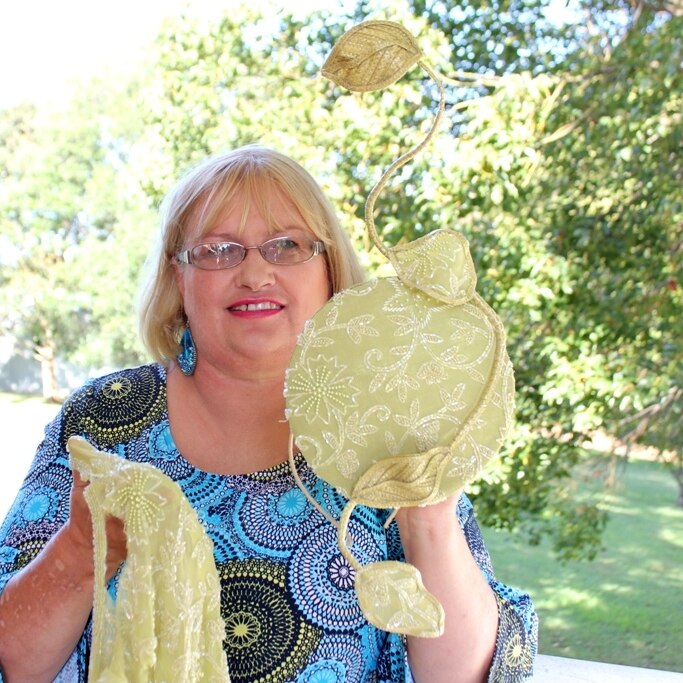 Milliner Caren Stevenson stands holding beaded material and a hat which has been made out of the material