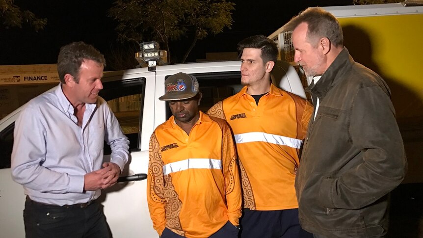 The two politicians talk to two community night patrol officers. All four people lean on a van.