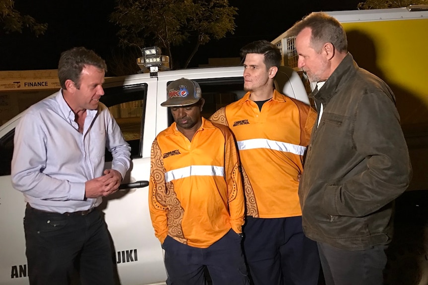 The two politicians talk to two community night patrol officers. All four people lean on a van.