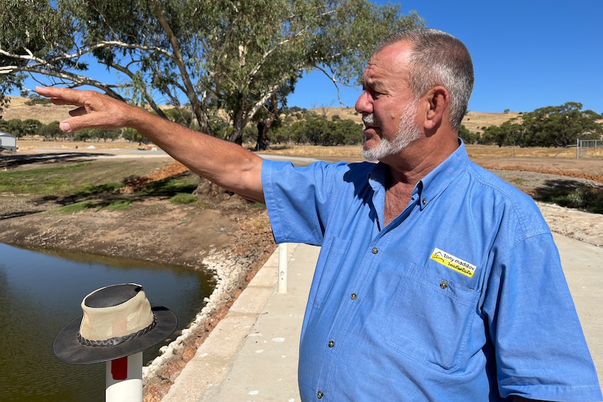 A middle-aged man with short, silvery hair and a neat white beard stands on a country property, pointing at something.