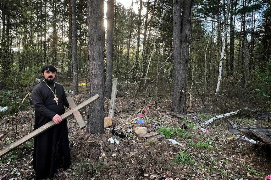 A priest stands in a forest holding a large wooden cross. 