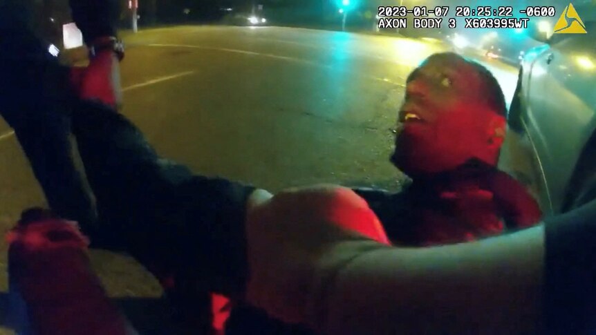 A screenshot shows a young black man looking up at police as he is violently arrested.