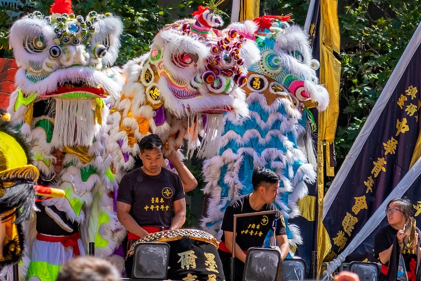Two male drummers and a female cymbalist perform with Lion dancers behind them.