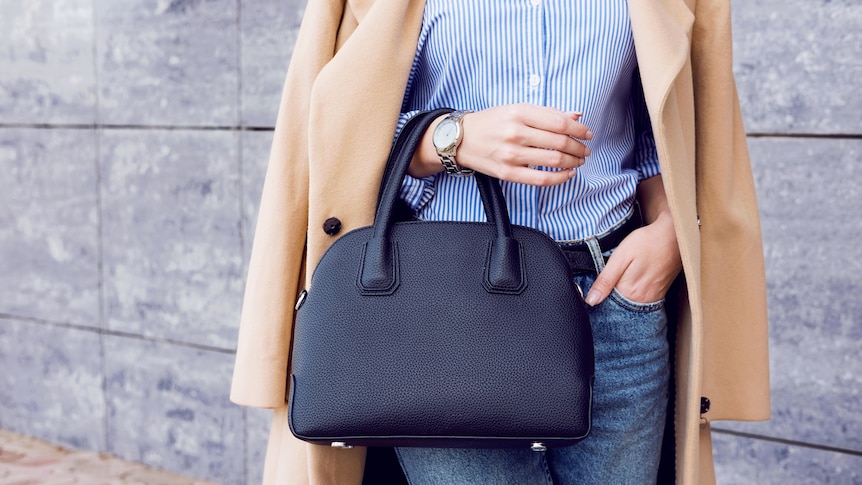 A stylish woman in a beige coat and jeans holds a large black handbag.