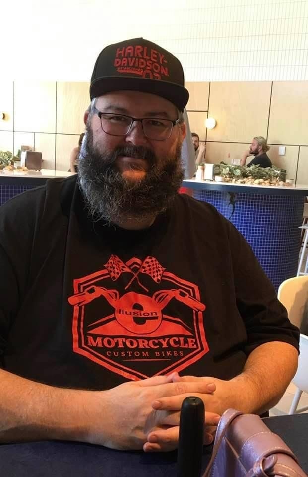A man with a beard smiles, wearing a hat that says Harley Davidson, sitting inside an eatery.