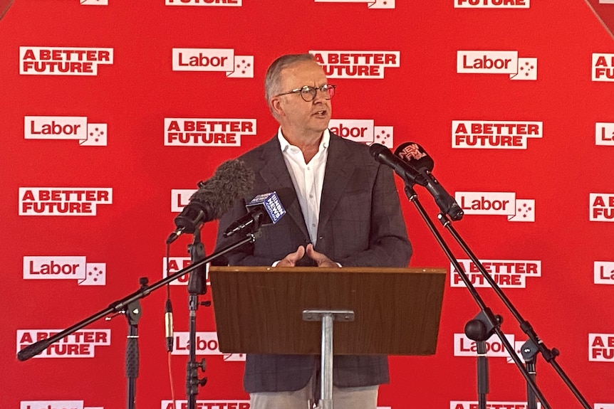 A man stands at a lectern surrounded by microphones in front of a red backdrop with Labor slogans.