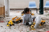 A little girl sits in a sandpit, surrounded by yellow toy dump trucks.