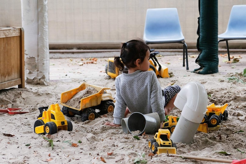 A little girl sits in a sandpit, surrounded by yellow toy dump trucks.